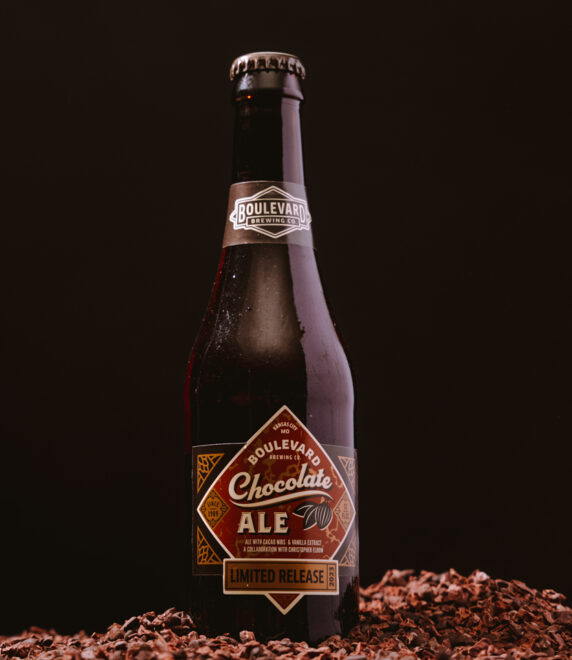 A bottle of Boulevard Chocolate Ale sits on a bed of cocoa nibs against a dark background. The label reads "Limited Release," highlighting the special nature of this beer. The setup emphasizes a rich chocolate theme.