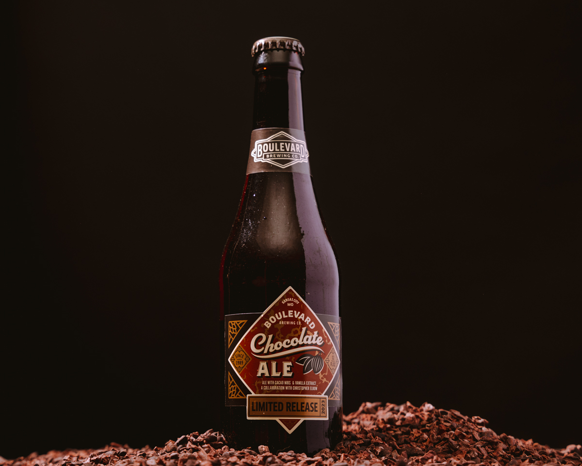 A bottle of Boulevard Chocolate Ale sits on a bed of cocoa nibs against a dark background. The label reads "Limited Release," highlighting the special nature of this beer. The setup emphasizes a rich chocolate theme.