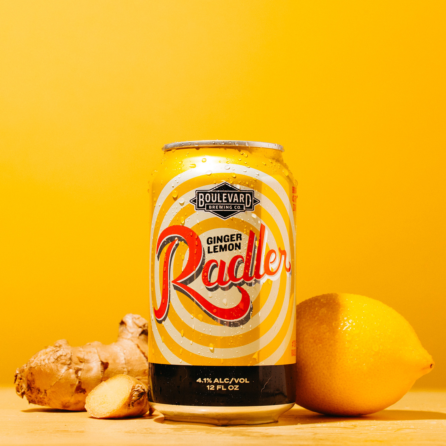 A can of Boulevard Brewing Co.'s Ginger Lemon Radler sits against a vibrant yellow background, alongside a whole lemon and a piece of ginger. The can features a spiral design with the text "Ginger Lemon Radler.