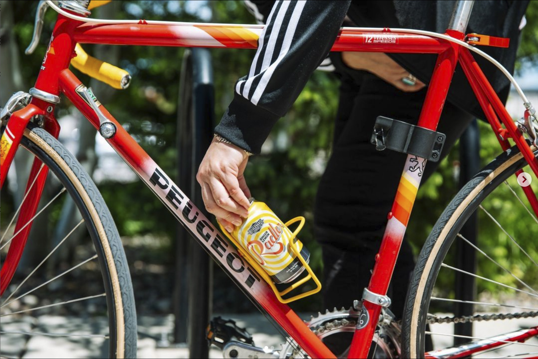 A person places a can of root beer into the yellow bottle holder of a red Peugeot bicycle. The bicycle features yellow and red details and is parked outdoors on a sunny day. The person is wearing a striped jacket and dark pants.