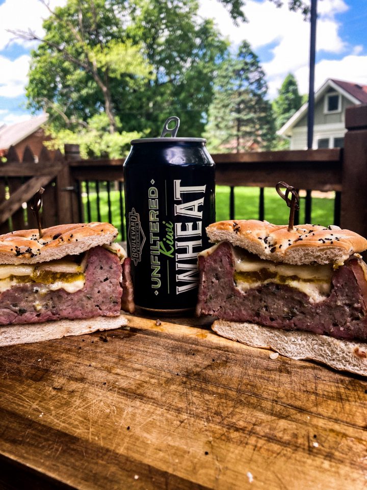 A large sandwich, cut in half, sits on a wooden surface with a can of unfiltered Kiwi Wheat beverage behind it. The sandwich has visible layers of meat and cheese, and is topped with seasoning. A lush garden is seen in the background.