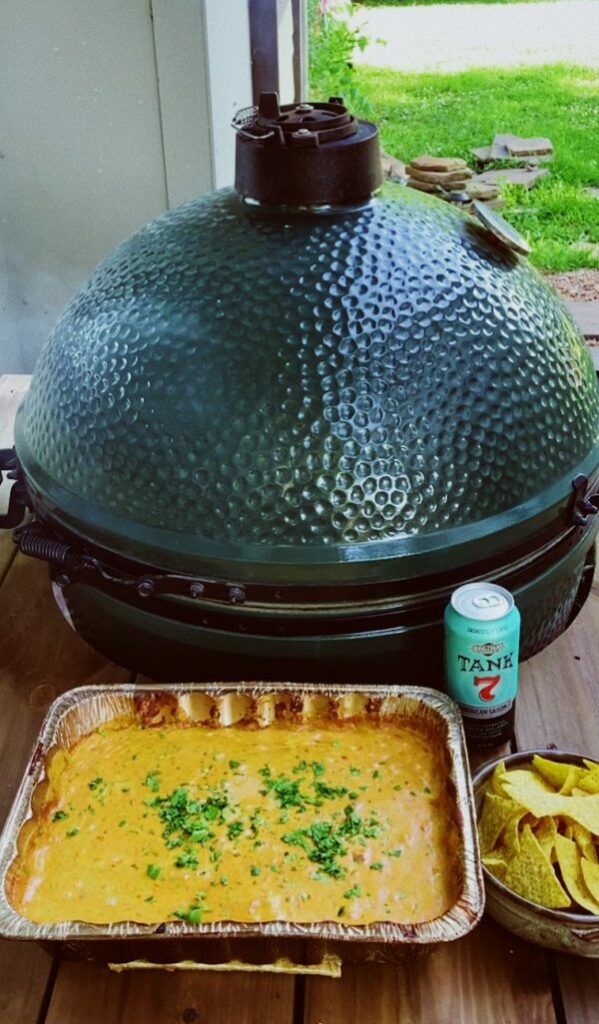 A green ceramic grill with a textured dome lid is on a wooden table. In front, there's a tray of cheesy dip garnished with herbs, a bowl of tortilla chips, and a can of Tank 7 beer. Green grass is visible in the background.