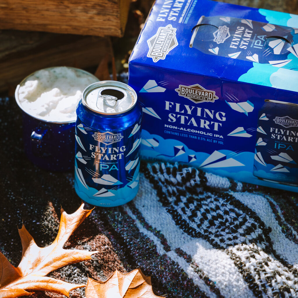 A can of Boulevard Brewing Co.'s Flying Start non-alcoholic IPA sits next to a blue mug filled with a frothy beverage. A 6-pack of Flying Start IPA is to the right on a patterned blanket. Autumn leaves are scattered, adding a seasonal touch to the outdoor scene.