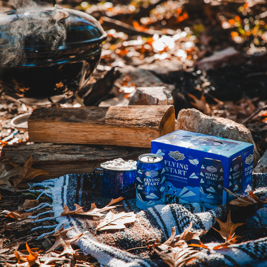 A cozy outdoor setup with a six-pack of Flying Start IPA beer, a cup with marshmallows, and a kettle on a striped blanket. Logs and autumn leaves are scattered around, and a grill is smoking in the background. Bright sunlight filters through the trees.