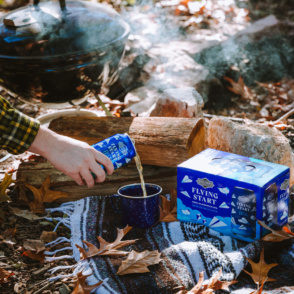 A person pours beer from a blue can labeled "Flying Start IPA" into a blue speckled camping mug. The scene is outdoors with autumn leaves on the ground, a striped blanket, a small pile of logs, and a smoking grill in the background next to a case of the same beer.