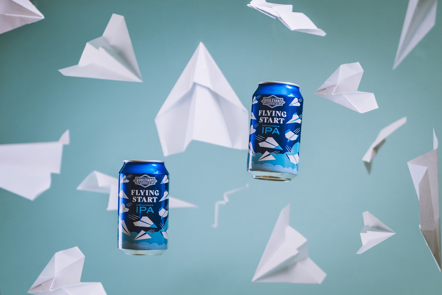 Two blue and silver cans of "Flying Start IPA" are suspended mid-air against a light blue background. Surrounding the cans are multiple white paper airplanes in various positions, giving a sense of motion and flight.