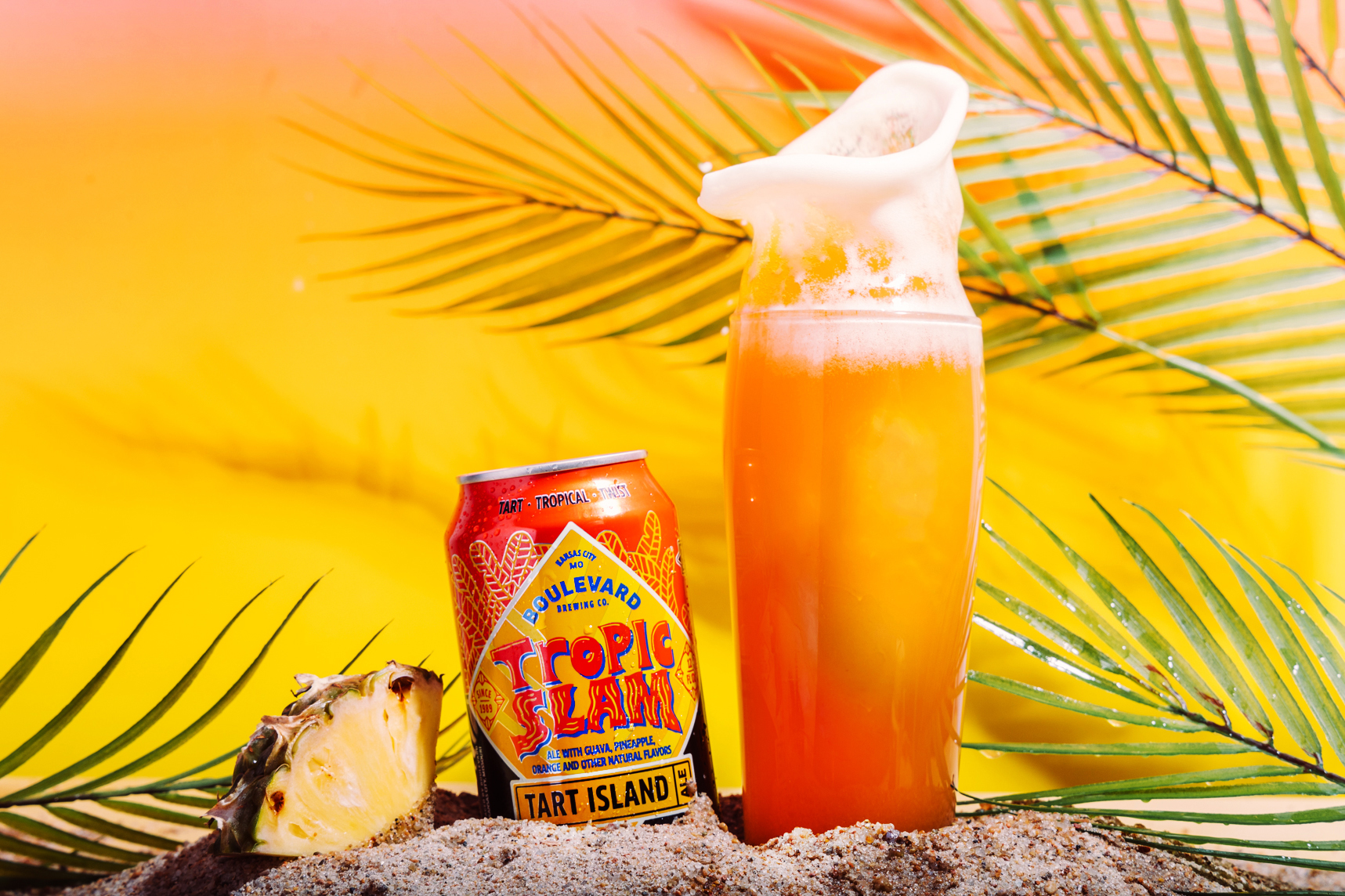 A can of Tropic Slam Tart Island Ale is placed next to a tropical-themed drink in a tall glass, filled with frothy orange liquid. Both sit on sandy ground, surrounded by palm leaves and a pineapple slice, against a warm yellow-orange background.