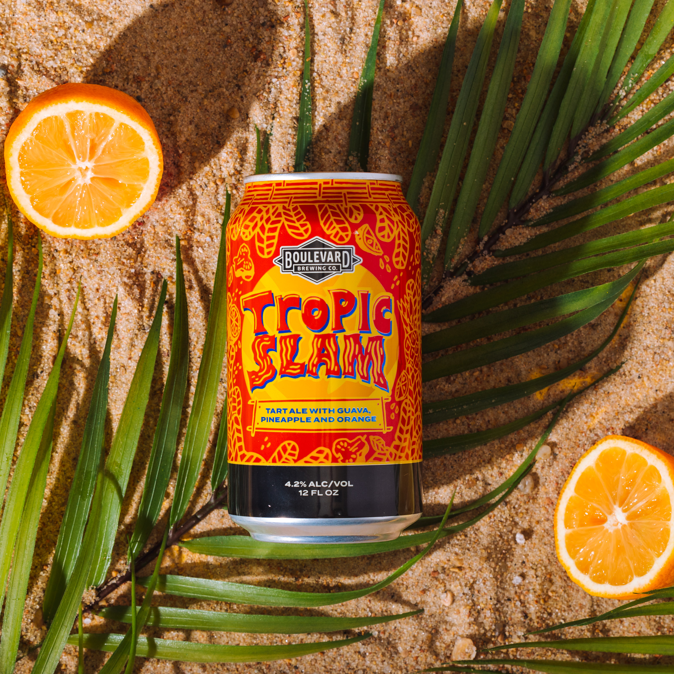 A can of Boulevard Brewing Co.'s Tropic Slam tart ale sits on sandy ground, surrounded by green palm leaves and orange slices. The can features vibrant orange and yellow tropical designs and highlights flavors of guava, pineapple, and orange.