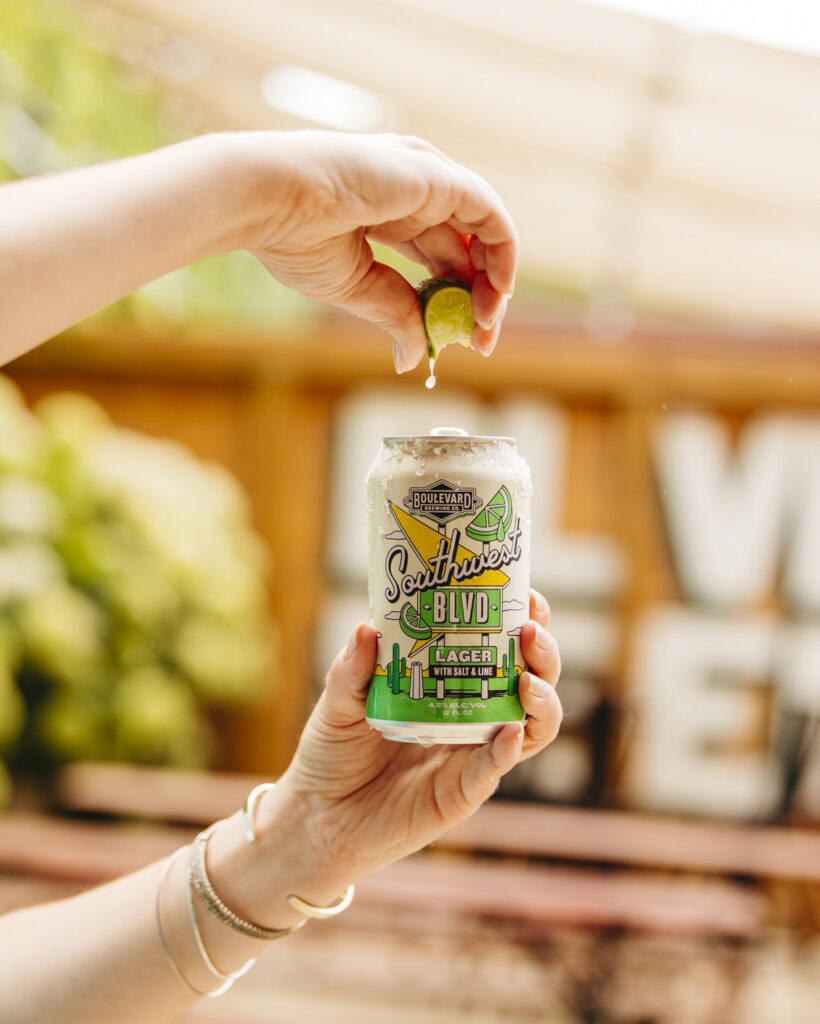 A person squeezes a slice of lime over an open can of Boulevard Brewing Company's Southwest BLVD Lager. The background features blurred outdoor furniture and greenery. The can design includes a breezy Southwest motif with cacti and sun rays.