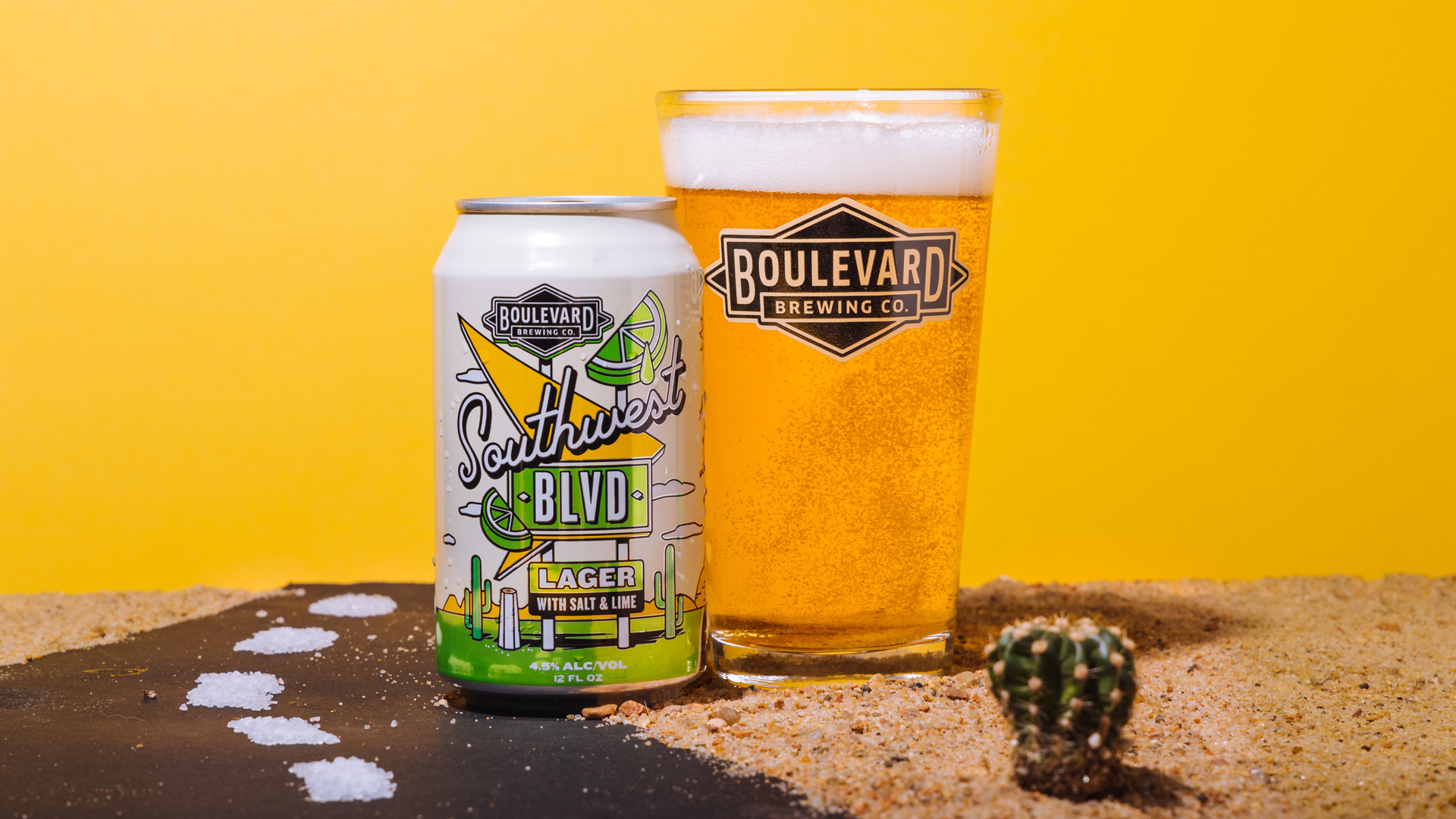 A can of Boulevard Brewing Co. Southwest BLVD Lager with Salt & Lime sits beside a filled pint glass against a yellow background. The setup includes sand, a small cactus, and a road leading up to the can, evoking a desert scene.