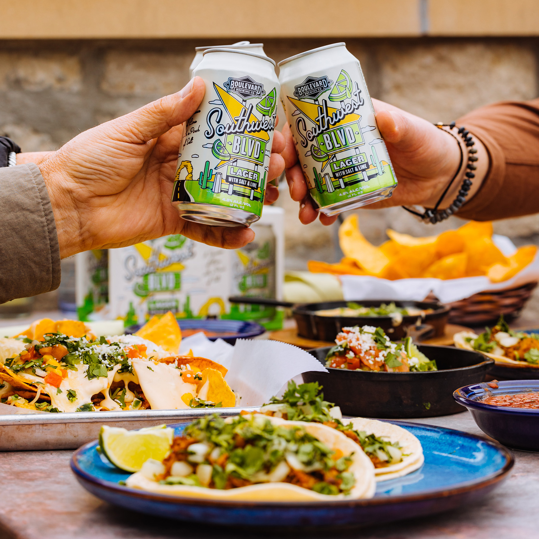 Two people clink cans of Southwest Blvd beer over a table filled with tacos, nachos, and various Mexican dishes, with colorful ingredients like cilantro, cheese, and limes. The scene appears to be a lively outdoor meal.