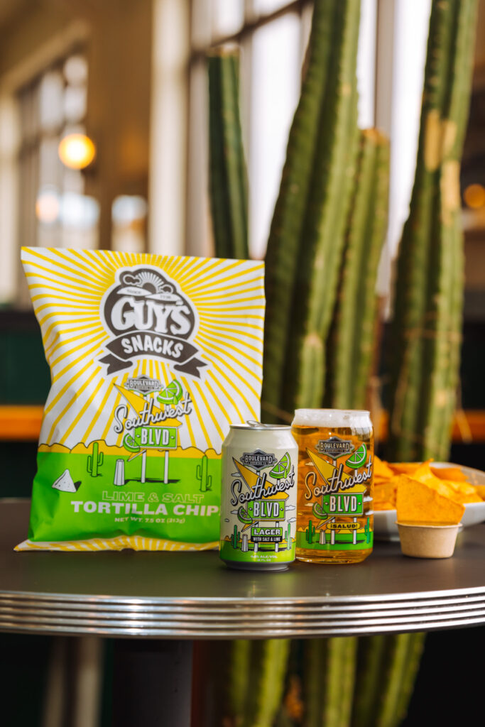 A table with a bright green and yellow bag of Guy's Snacks lime & salt tortilla chips, a can and a glass of Southwest Blvd lager beer, and a bowl of tortilla chips. A tall cactus is in the background.