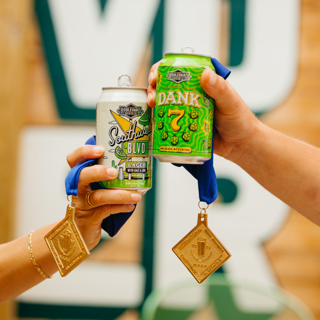 Two hands holding up Boulevard Brewing Co. beer cans - one a Southwest Blvd Lager and the other a Dank 7 IPA. Both cans have medals attached, with blue ribbons and gold plaques, against a blurred background.