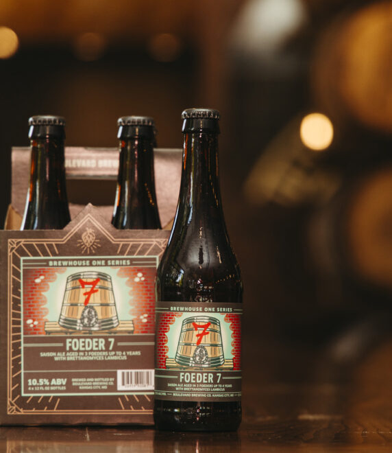 A close-up shot of 4-pack beer bottles, with one bottle placed in front. The labels on the bottles read "Foeder 7," part of the "Brew House One Series." Wooden barrels are blurred in the background. The beer has an alcohol content of 10.5% ABV.