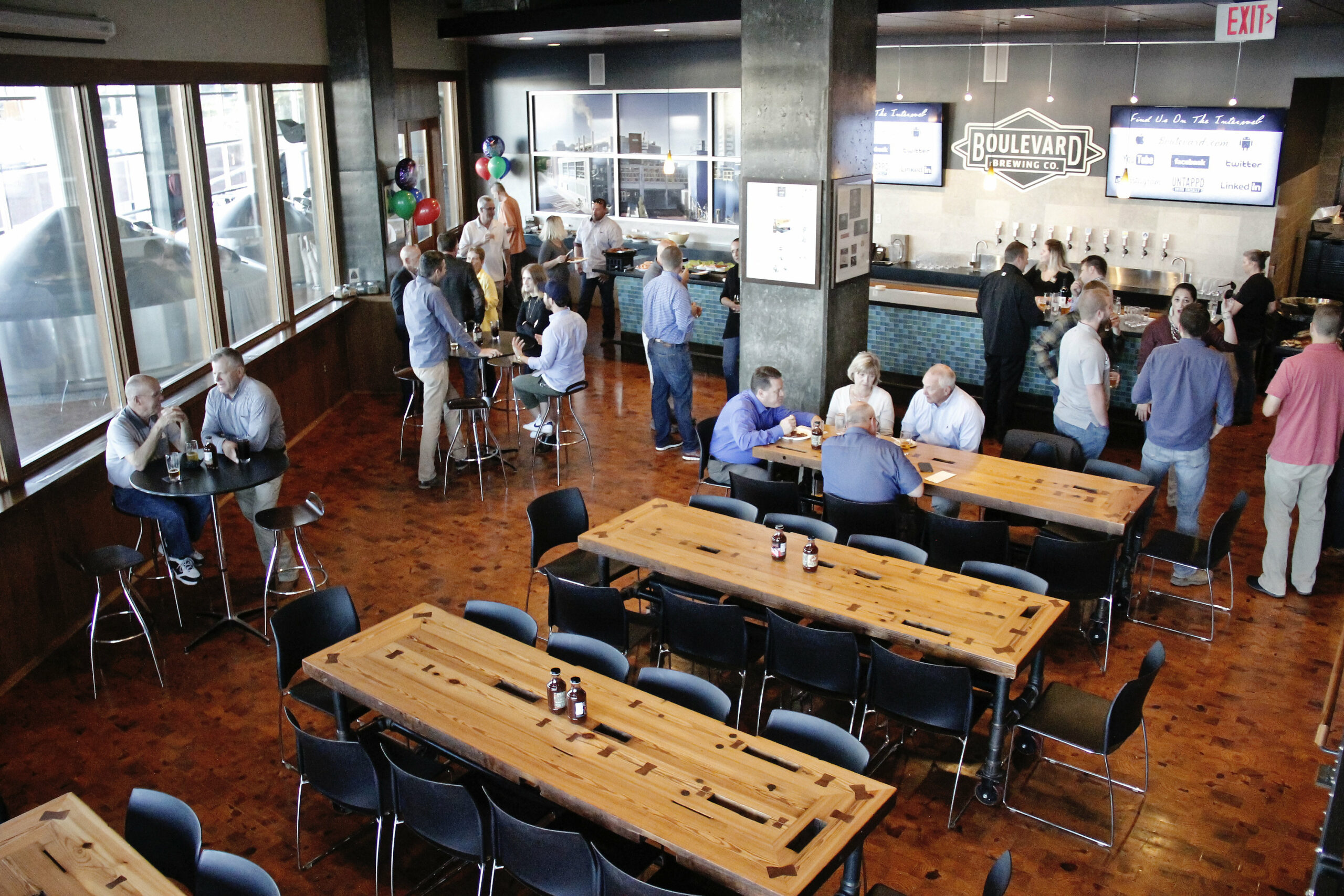 A modern brewery bar with high wooden tables and chairs. People are casually gathered in small groups, some seated and some standing, chatting and enjoying drinks. A bar with taps and a "Boulevard Brewing Co." sign is visible in the background.