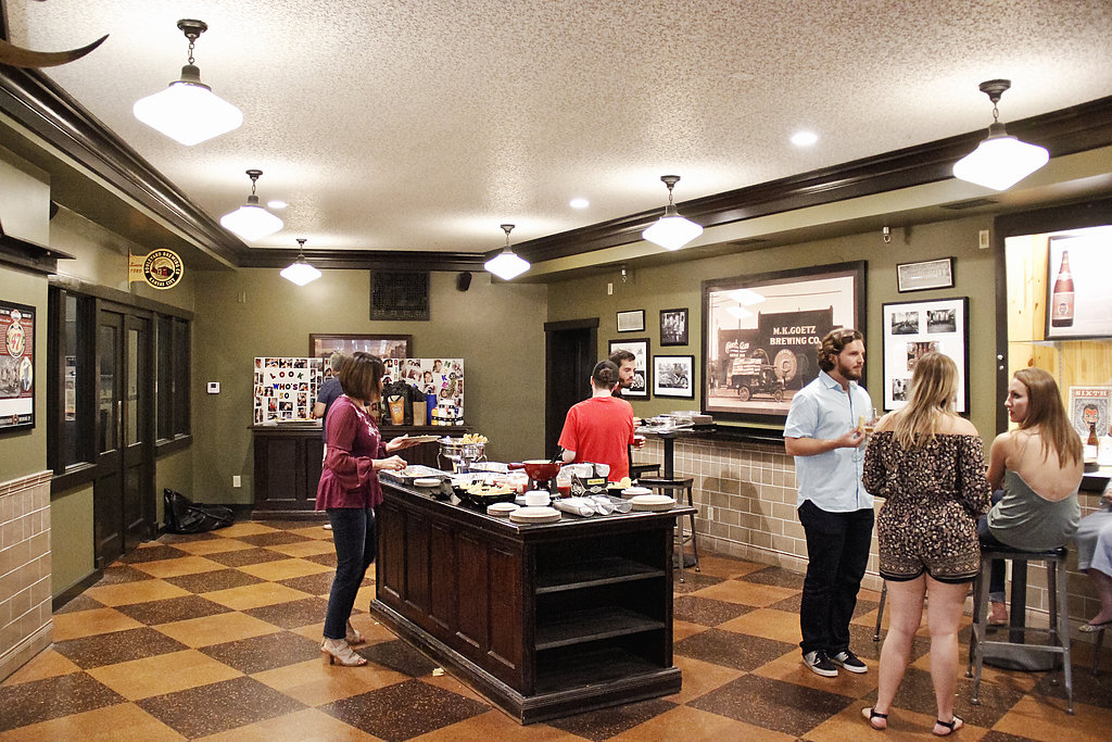 A warmly lit room features a buffet table with food and drink. Five people are conversing and enjoying the meal. The walls are adorned with framed pictures and vintage decor. The space has checkered flooring and high ceilings with hanging lights.