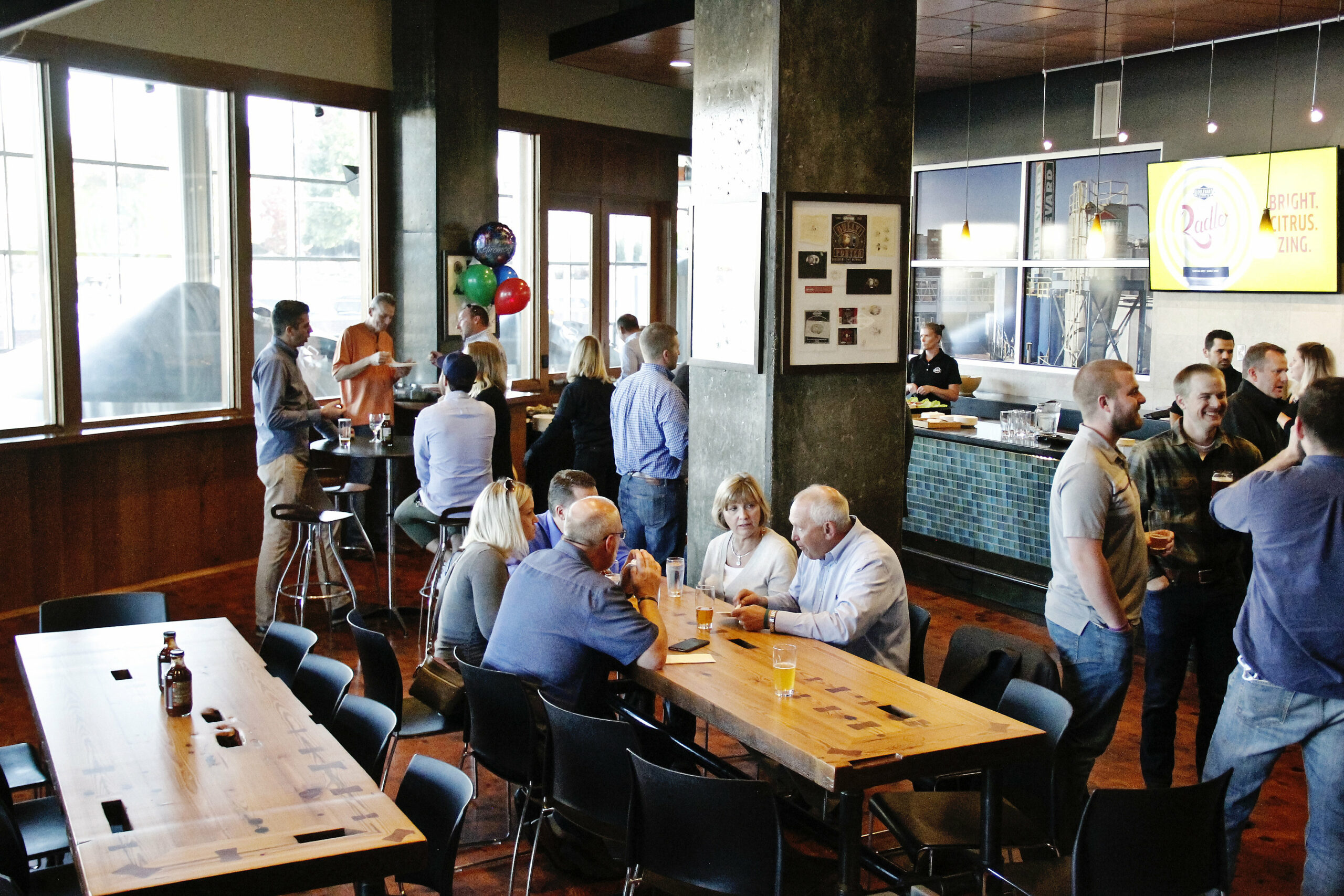 A group of people socializing in a modern, well-lit bar or restaurant. Some are standing and mingling near the bar area, while others are seated at tables, engaging in conversation. There are festive balloons in one corner, and a bartender is serving drinks.