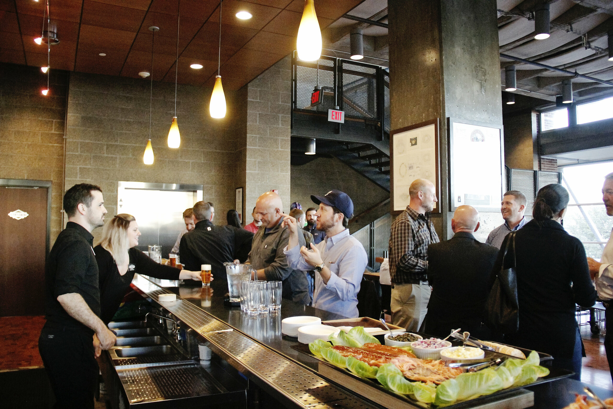 A lively bar scene with people engaged in conversations and enjoying drinks. In the foreground, various food items are laid out on the counter. The setting has modern decor with hanging lights, large windows, and a staircase in the background.