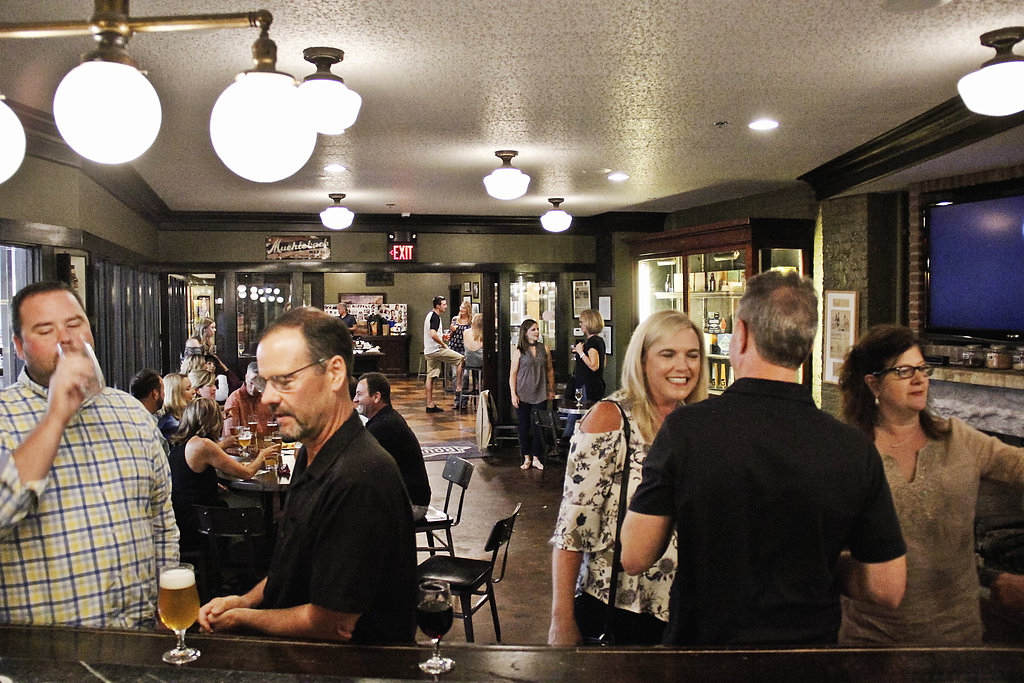 A lively pub scene with several people enjoying drinks and conversation. Most are standing, and a few are seated at tables. The pub is warmly lit with overhead lights, and a TV is mounted on the wall. The atmosphere appears casual and friendly.