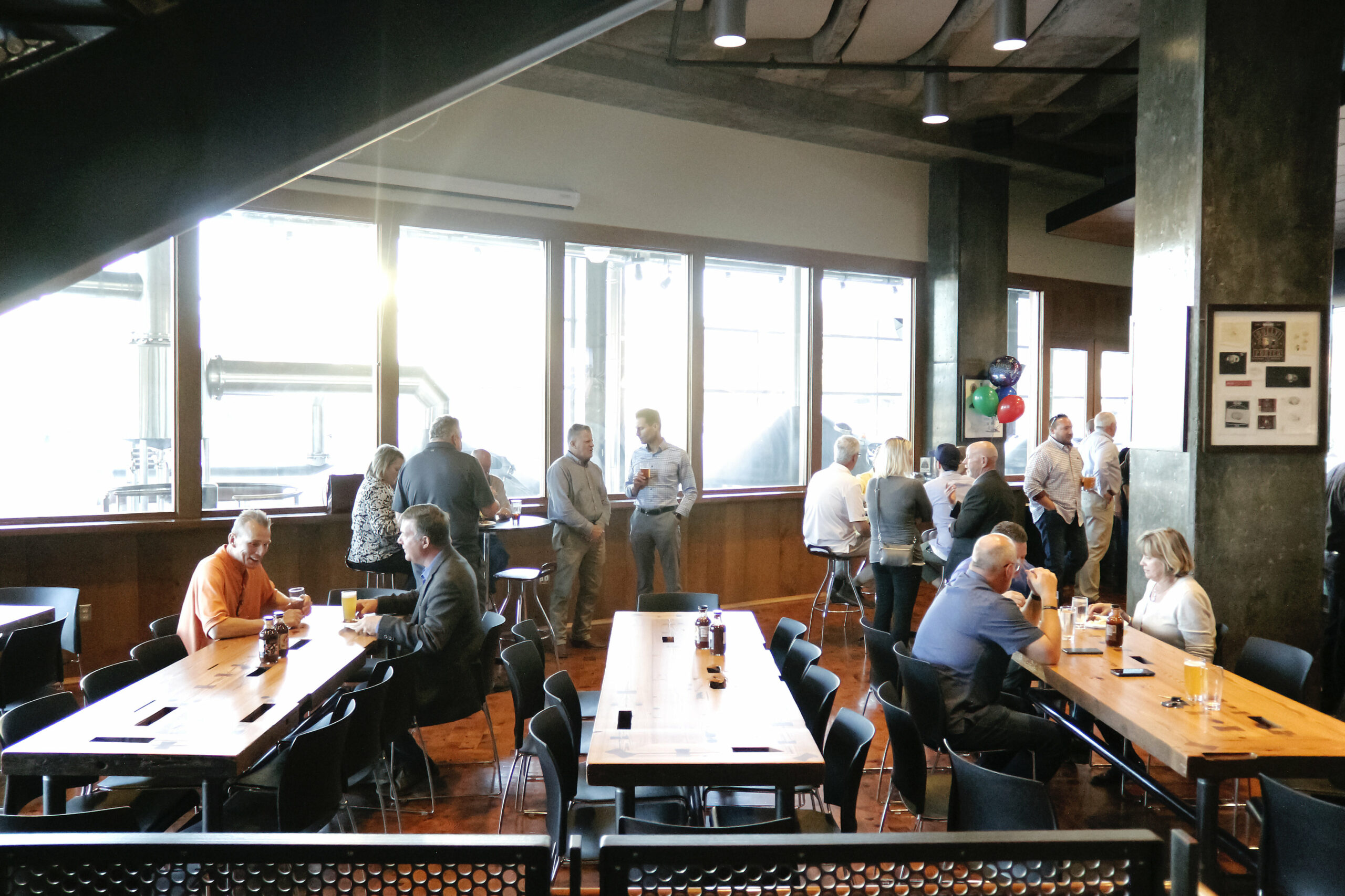 A group of people are gathered in a modern, spacious room with large windows. They are standing and sitting at wooden tables, engaging in conversation. The setting appears to be casual and social, with natural light streaming in through the windows.