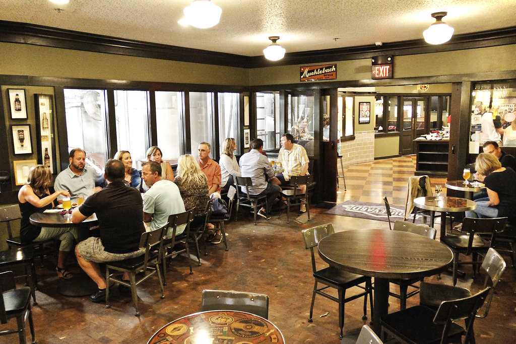 A cozy indoor brewery scene with multiple groups of people seated at tables, enjoying drinks and conversation. The room has a warm, inviting atmosphere with a mix of wooden and metal furniture and a view into the brewing area through large windows.