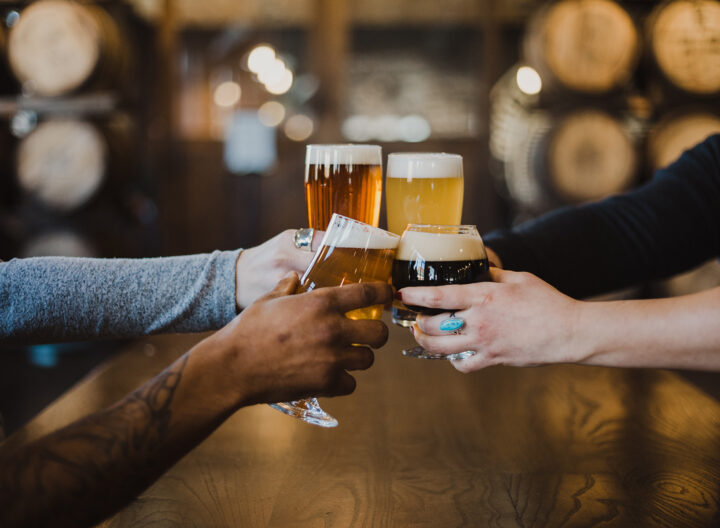 Four people clink glasses filled with various types of beer over a wooden table. In the background, large barrels are visible, giving a cozy, rustic atmosphere.