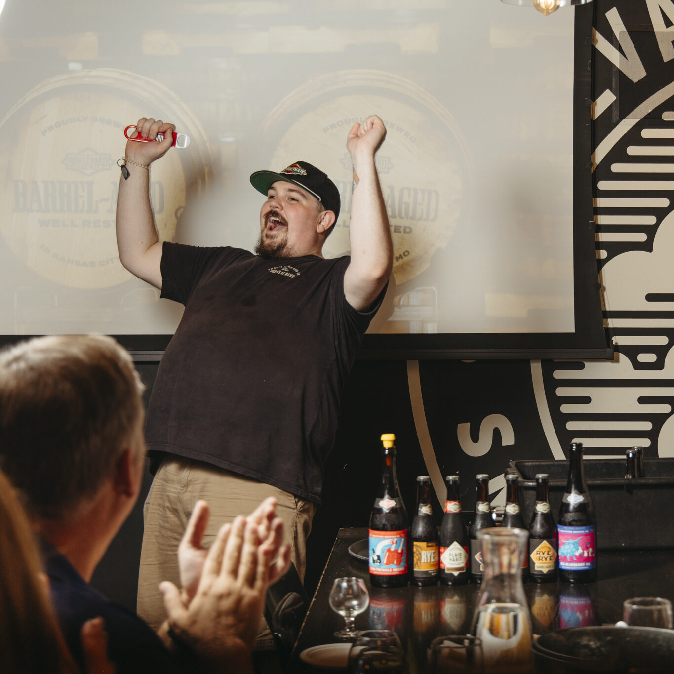 A man in a black shirt smiling with his hands in the air. In front of him are several beer bottles and happy guests.