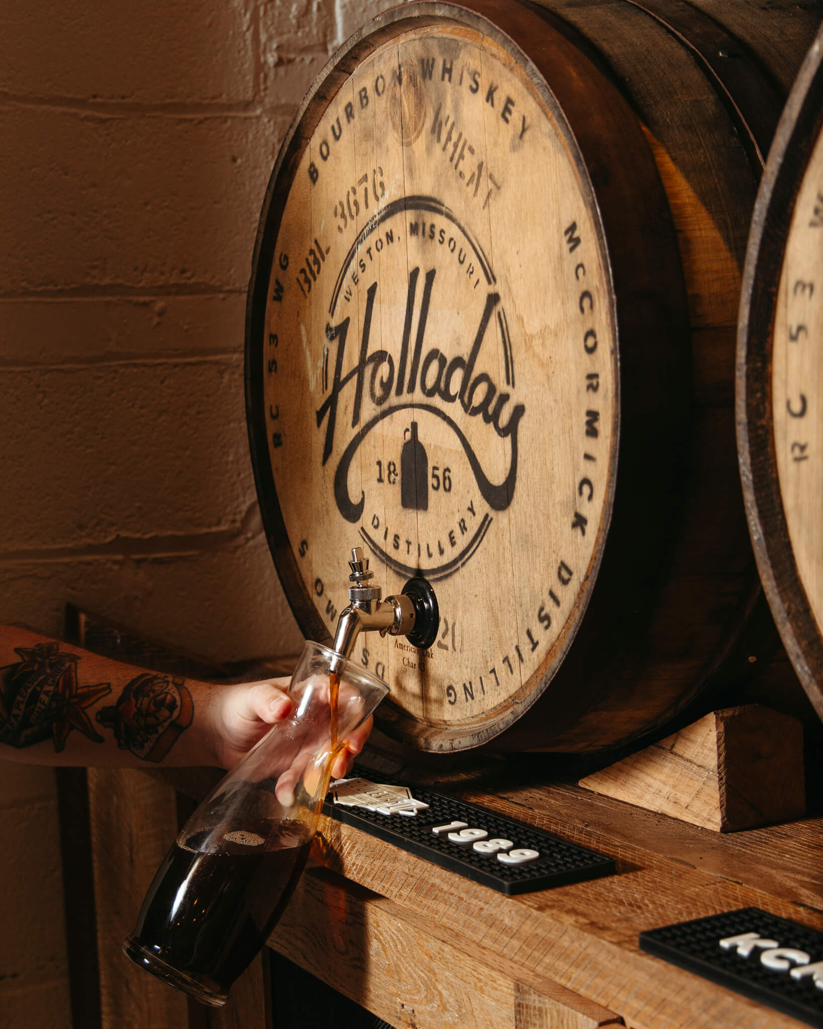 A person fills a glass pitcher with whiskey from a wooden barrel labeled "Holladay Distillery." The barrel, marked with "Bourbon Whiskey Wheat" and other inscriptions, is mounted on a wooden rack. The person's arm has visible tattoos.