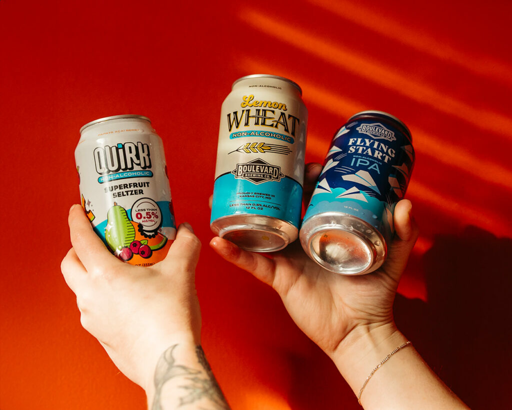 Two hands holding three beverage cans against a vibrant red background. Cans from left to right: Quirk Superfruit Seltzer, Lemon Wheat Non-Alcoholic Beer, and Flying Start IPA. Sunlight casts soft shadows on the wall.