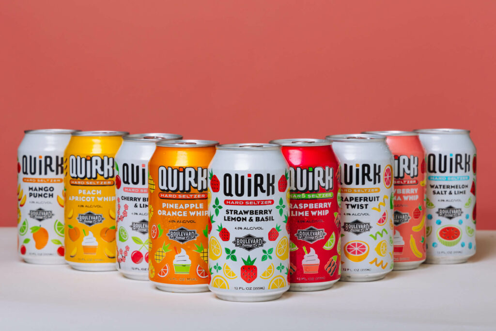 A display of twelve Quirk Hard Seltzer cans arranged in two rows against a peach-colored background. The cans feature various flavors including mango punch, pineapple orange whip, raspberry lime whip, and grapefruit twist, with colorful and vibrant designs.