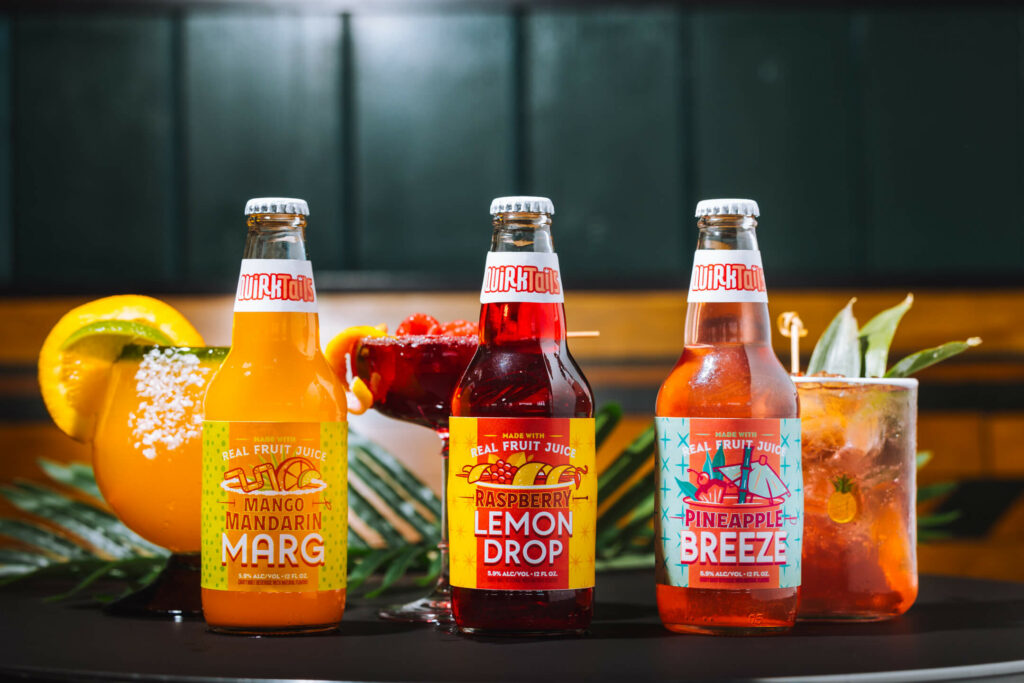 Three bottles of fruity soda on a table, labeled Mango Mandarin Marg, Raspberry Lemon Drop, and Pineapple Breeze. Four colorful cocktails are arranged behind and around the bottles, garnished with fruit and herbs. Background features a dark green wall.