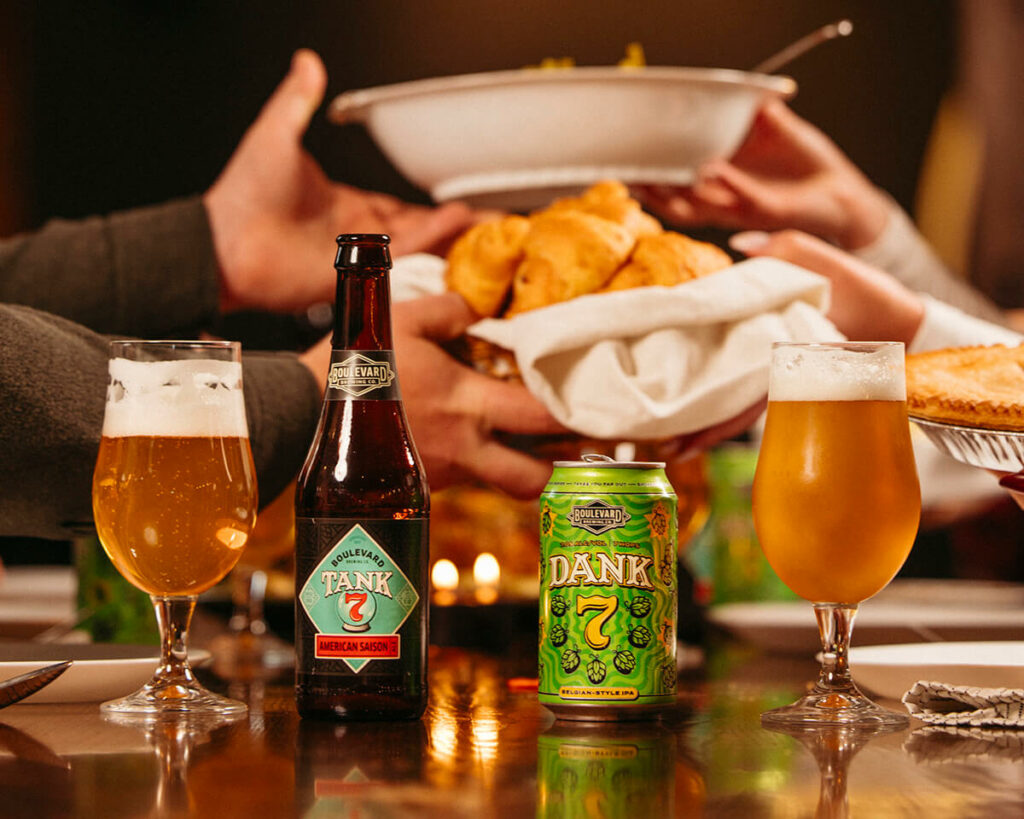 A bottle and can of Tank 7 beer are on a table along with two filled beer glasses. People in the background are passing a bowl of bread rolls across the table, creating a warm and inviting dining scene.