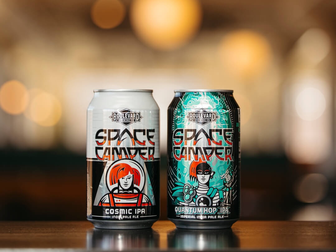 Two cans of Boulevard Brewing Co.'s Space Camper IPA are displayed on a wooden surface. The left can features "Cosmic IPA" with an illustration of an astronaut in red, while the right can showcases "Quantum Hop IPA" with an astronaut in green. The background is blurred.