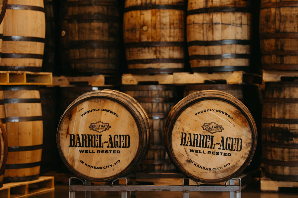 A stack of wooden barrels is arranged in rows, with two prominent barrels in the foreground labeled "Barrel-Aged Well Rested" and "Proudly Brewed in Kansas City, MO." The barrels appear aged and are part of a brewery or distillery setup.