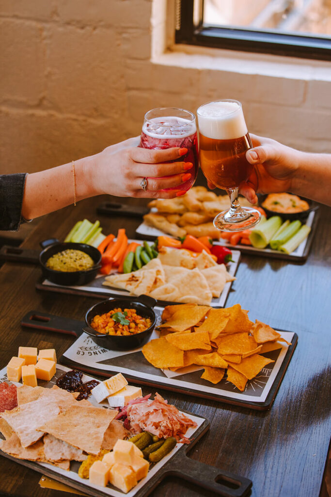 Two people clink glasses over a table filled with a variety of appetizers. The table has cheese, crackers, chips, dip, cut vegetables, hummus, and other snacks. The setting is indoors, with a brick wall and window in the background.