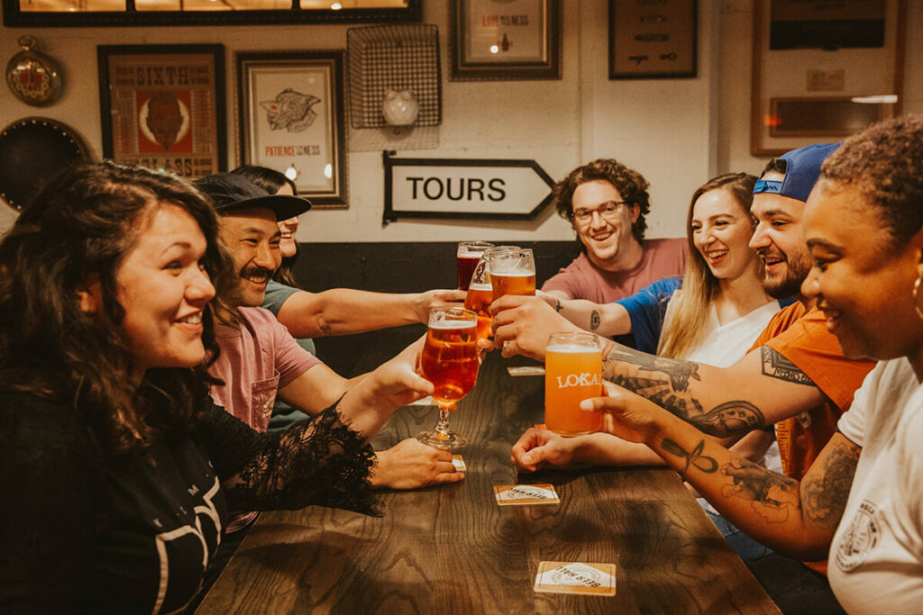 A diverse group of six friends sits around a wooden table at a bar, smiling and clinking their beer glasses in a toast. The background features various wall decorations, including a sign pointing to "Tours.