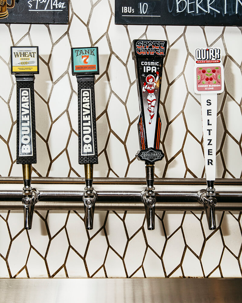 Four beverage taps with labeled handles are lined up against a tiled backsplash. The handles display: "Wheat" by Boulevard, "Tank 7" by Boulevard, "Space Camper Cosmic IPA," and "Quirk Seltzer." A price board is partially visible above the taps.