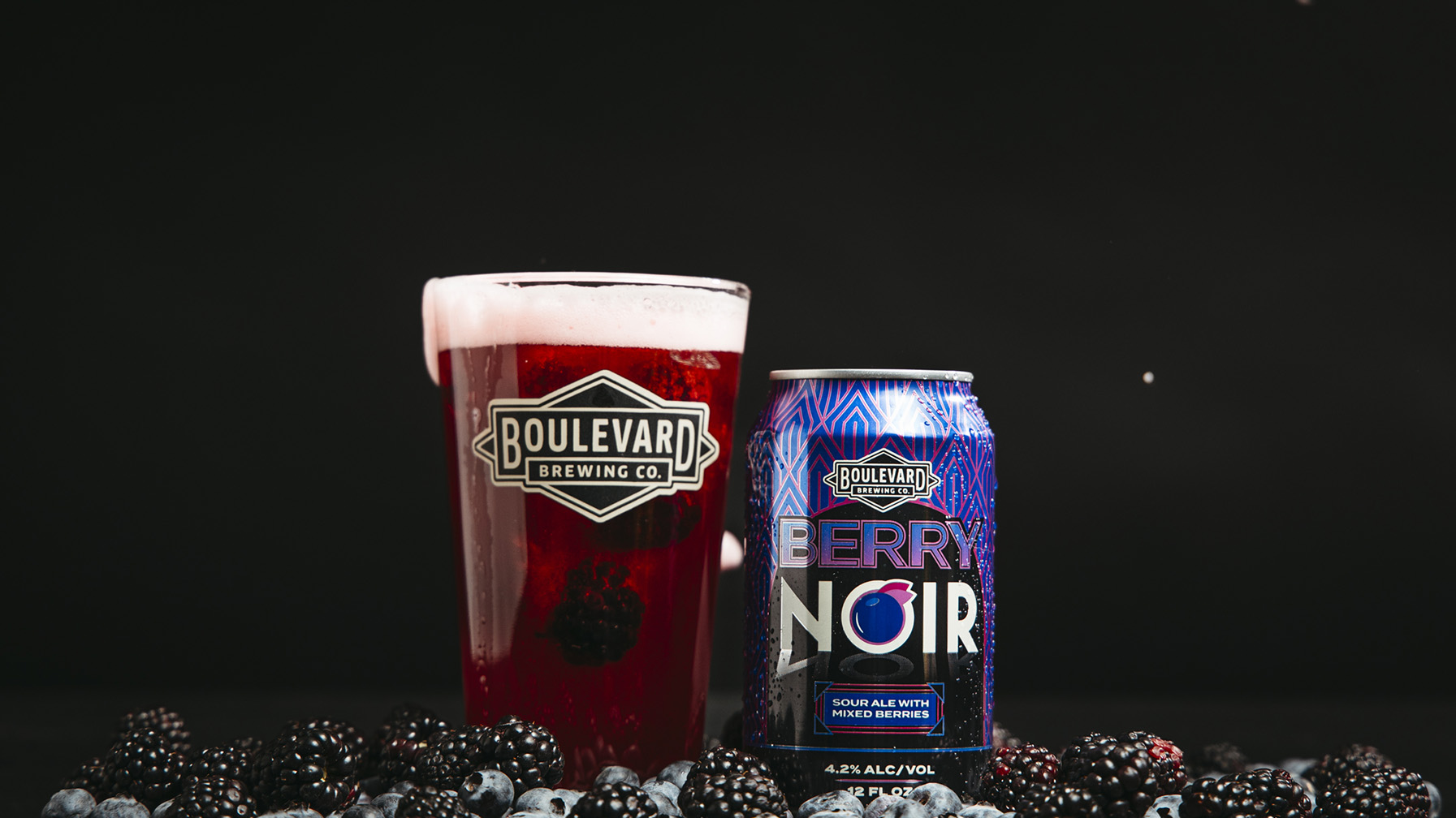 A glass filled with a deep red beverage labeled "Boulevard Brewing Co." stands beside a blue can labeled "Berry Noir." Surrounding them are scattered blackberries and blueberries against a dark background.