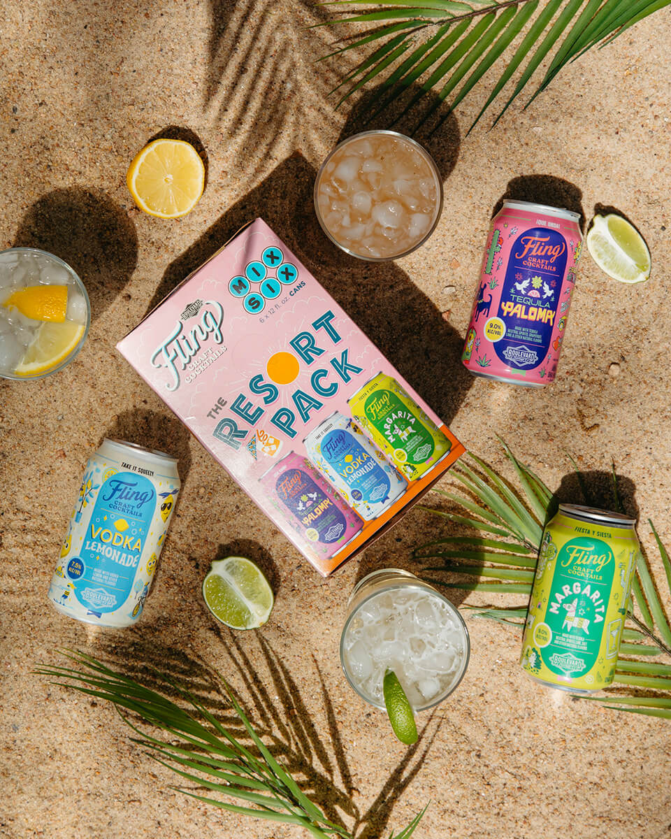 A box labeled "The Resort Pack" by Fling Craft Cocktails is surrounded by four cans of different cocktails on a sandy beach with palm leaves. Two glasses filled with ice and drinks, along with lime wedges, are also placed around the box and cans.