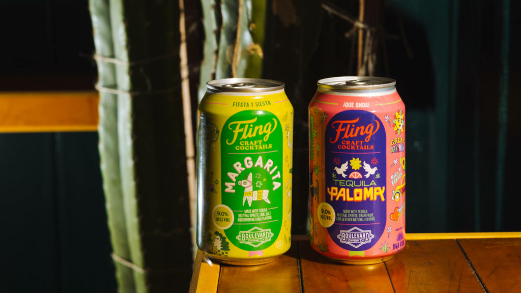 Two colorful cans of Fling Craft Cocktails are positioned in front of a cactus plant. The can on the left is a Margarita flavor, green with yellow accents. The can on the right is a Tequila Paloma flavor, pink with orange accents. Both cans exhibit vibrant designs.