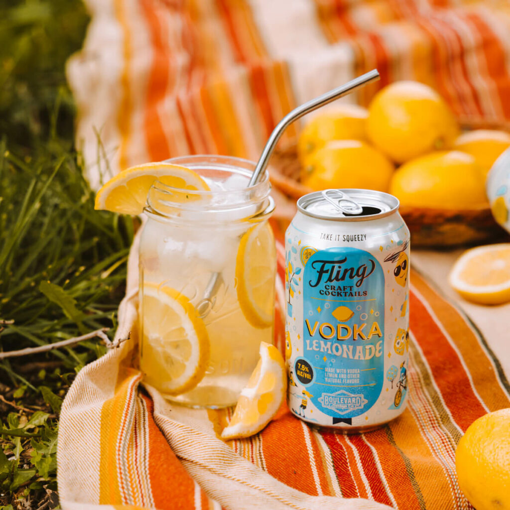 A can of Fling Craft Cocktails Vodka Lemonade is placed next to a glass mason jar filled with ice and lemon slices, topped with a metal straw. The setup is on a striped picnic blanket with whole lemons scattered around in the background.