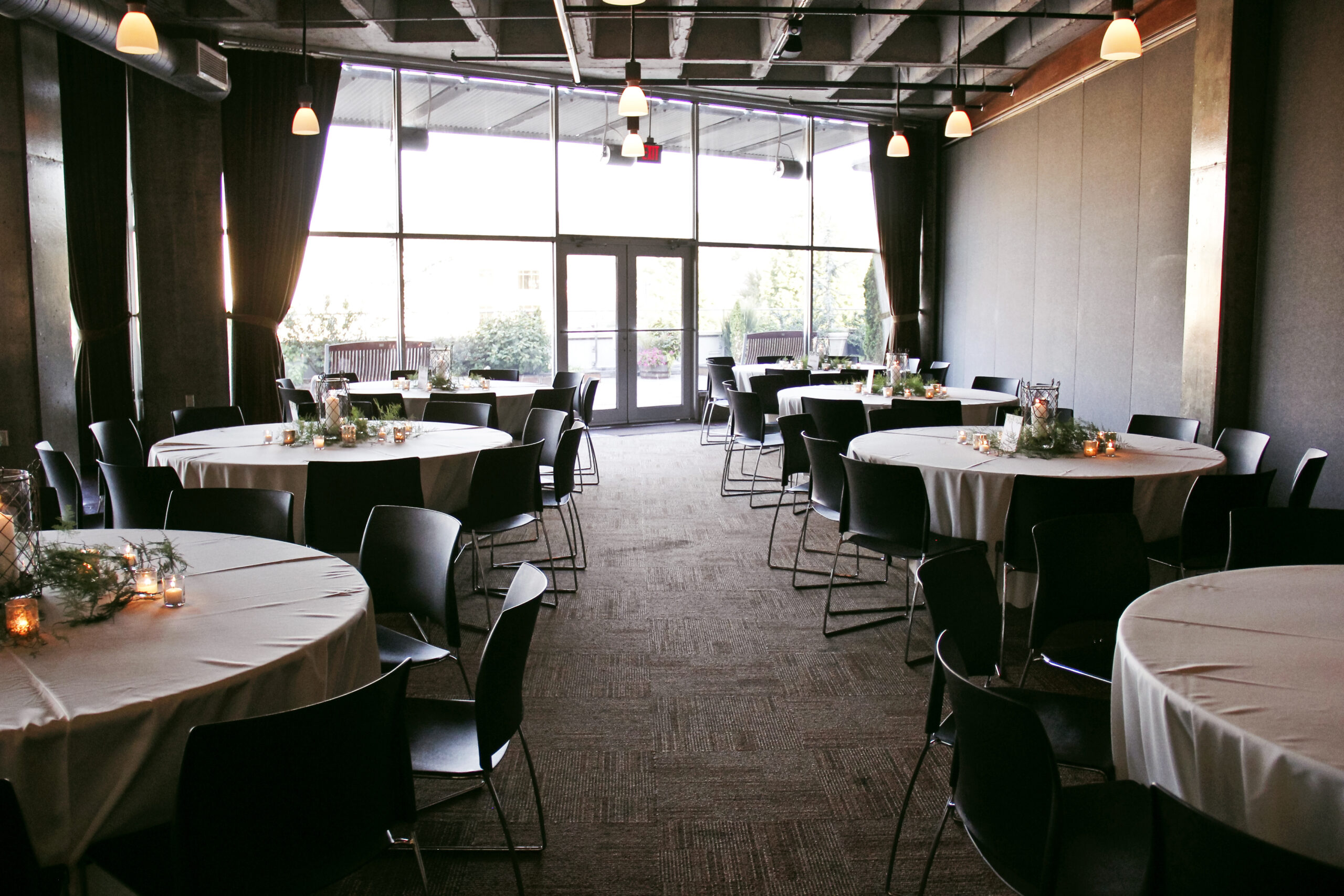 A decorated event space with round tables covered in white tablecloths and surrounded by black chairs. Each table has small centerpieces with candles and greenery. The room has large windows with natural light coming in and hanging pendant lights from a high ceiling.