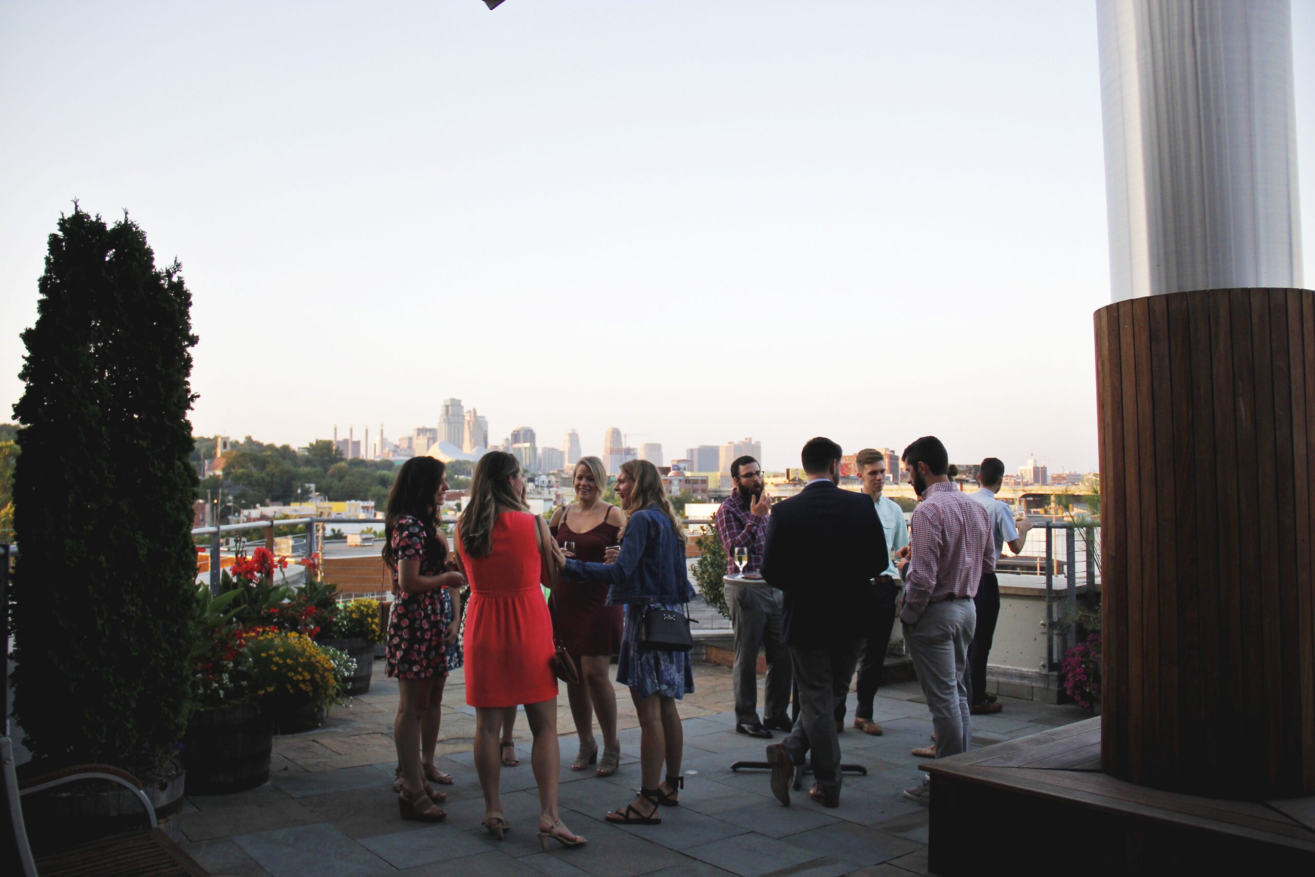 A group of people in casual and semi-formal attire socialize on a rooftop patio during the day. They stand in small clusters, holding drinks and engaging in conversation. The background shows a city skyline with various buildings under a clear sky.