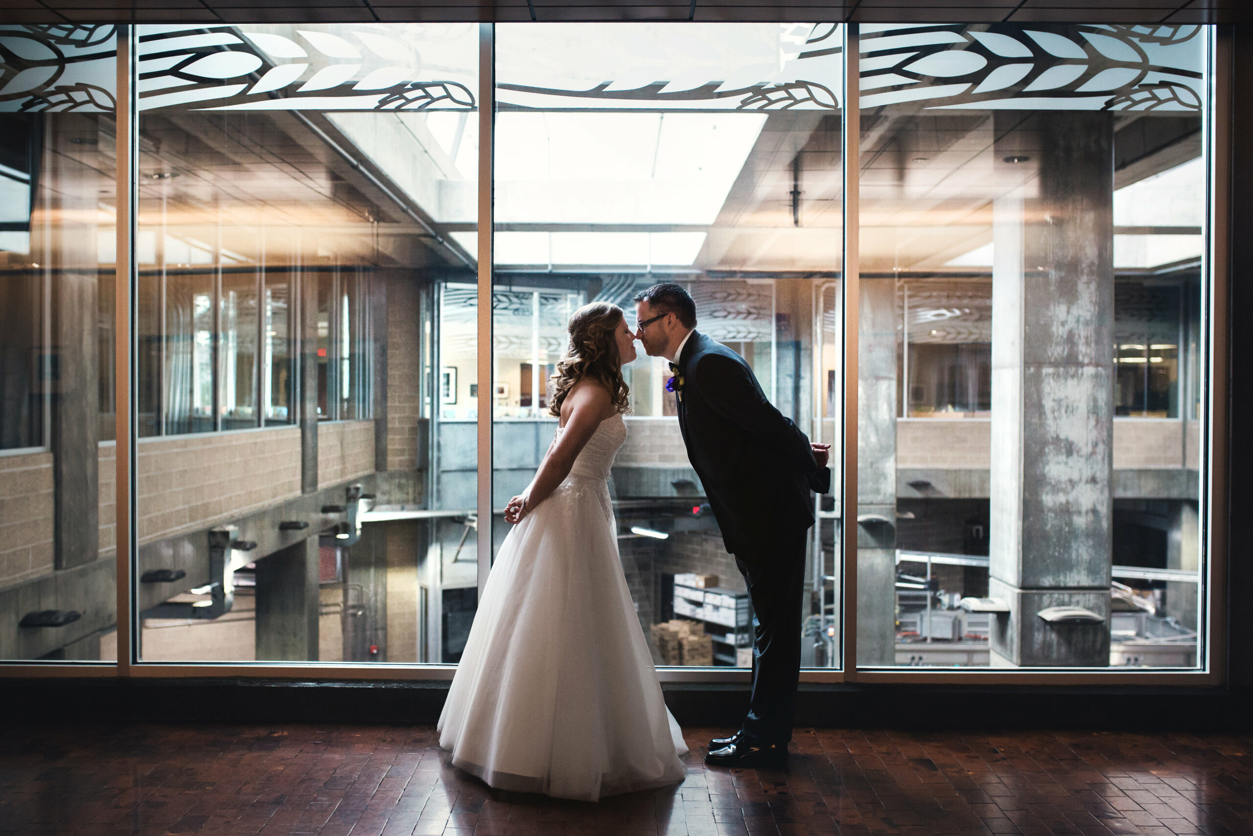 A couple is dressed in wedding attire, with the bride in a white gown and the groom in a black suit. They are standing indoors in front of large glass windows, leaning in for a kiss. The modern architecture behind them includes glass and concrete elements.