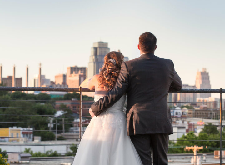 A couple dressed in wedding attire stands close together, facing away from the camera on a rooftop terrace. They gaze towards a cityscape with tall buildings in the distance, with the evening sky above. The groom has his arm around the bride's shoulders.