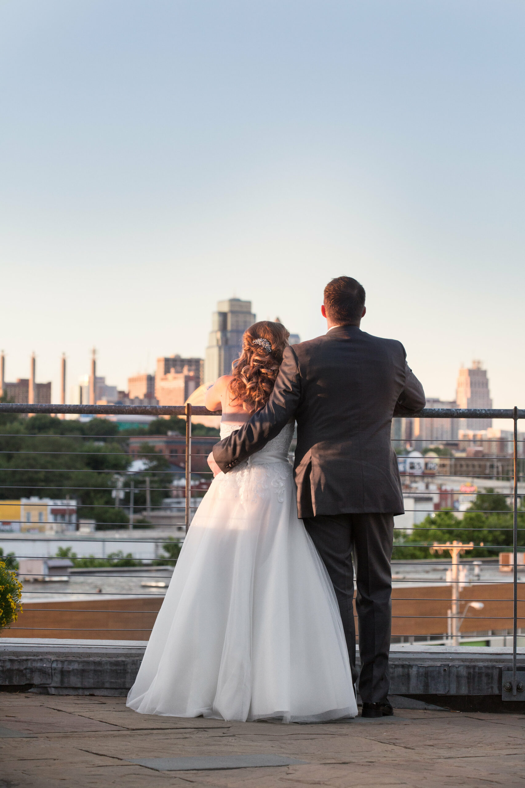 A couple dressed in wedding attire stands close together, facing away from the camera on a rooftop terrace. They gaze towards a cityscape with tall buildings in the distance, with the evening sky above. The groom has his arm around the bride's shoulders.