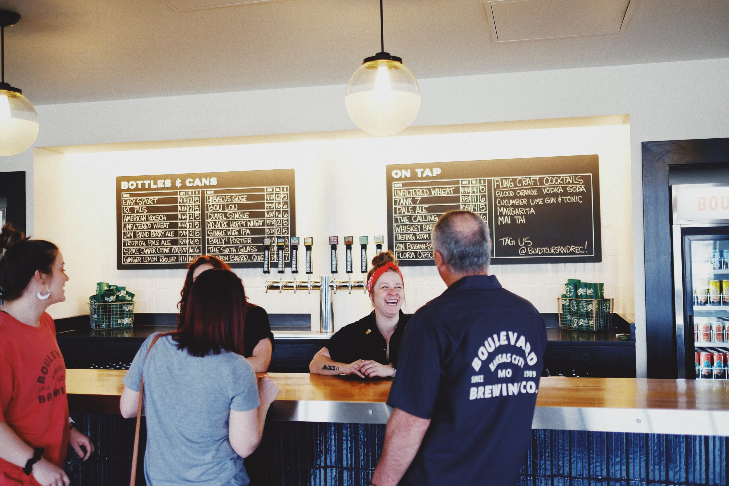 A group of people smiling and chatting at a bar. Behind the bar are beer taps and a menu board listing available beverages. The background includes hanging lights and a logo sign.