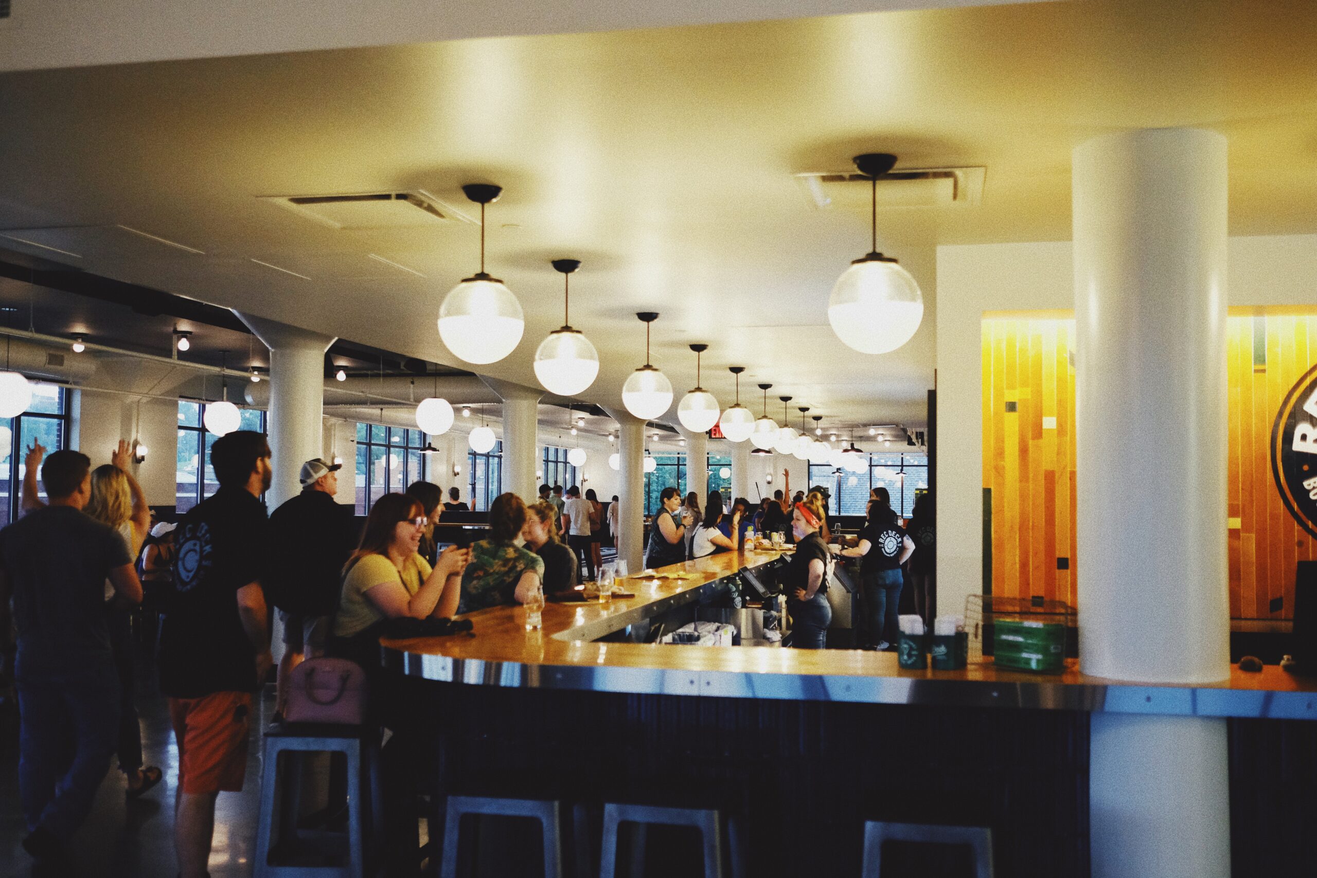 A lively bar scene with people gathered around a long, curved bar counter. The space features modern decor with round hanging lights, white columns, and a wooden accent wall. Patrons are engaged in conversations and enjoying drinks.