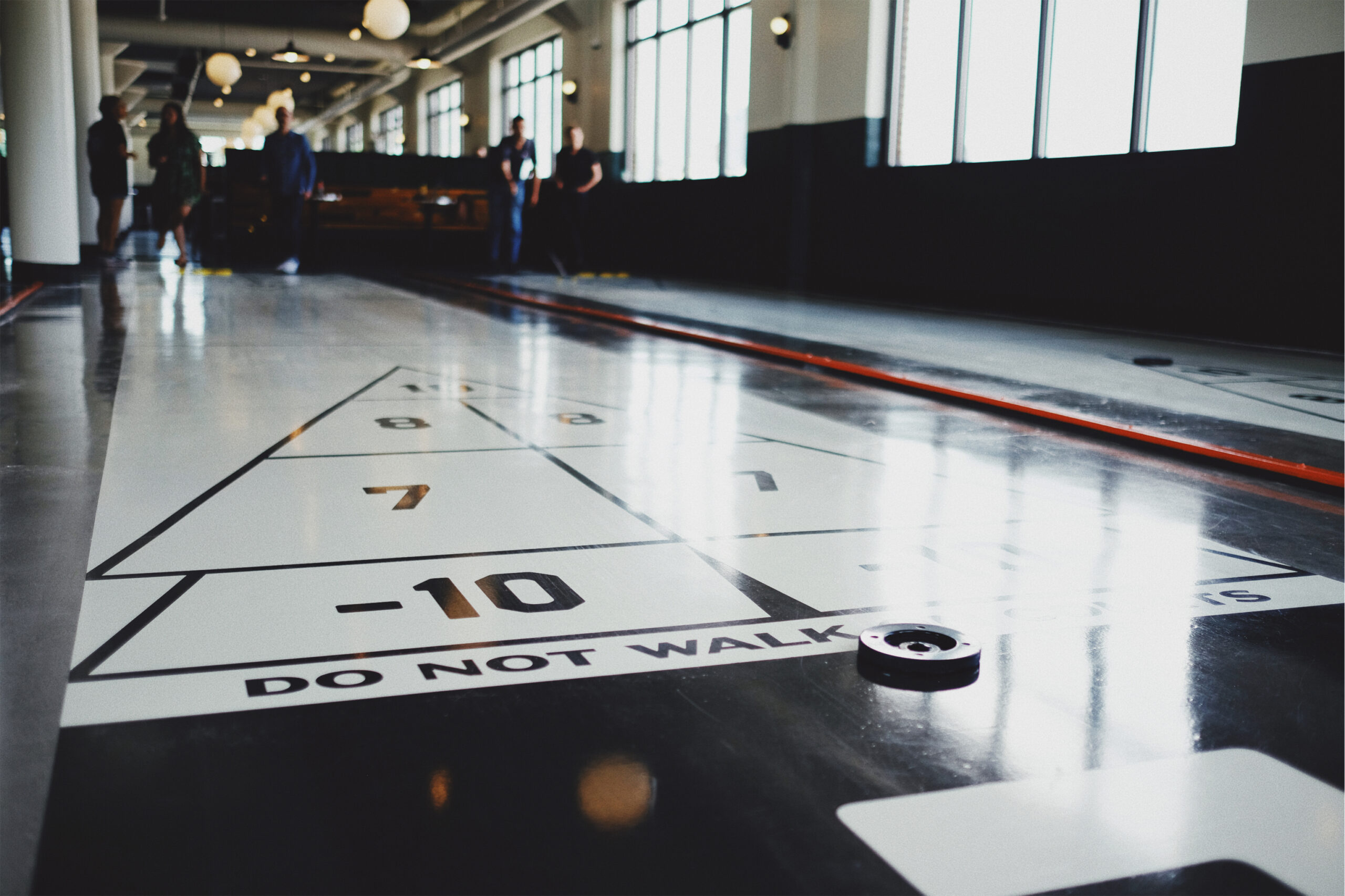 A shuffleboard table in an indoor recreational space, showing close-up of the scoring zones marked with numbers. There is a puck on the table, and several people are visible in the background, partially out of focus. Natural light enters through large windows.