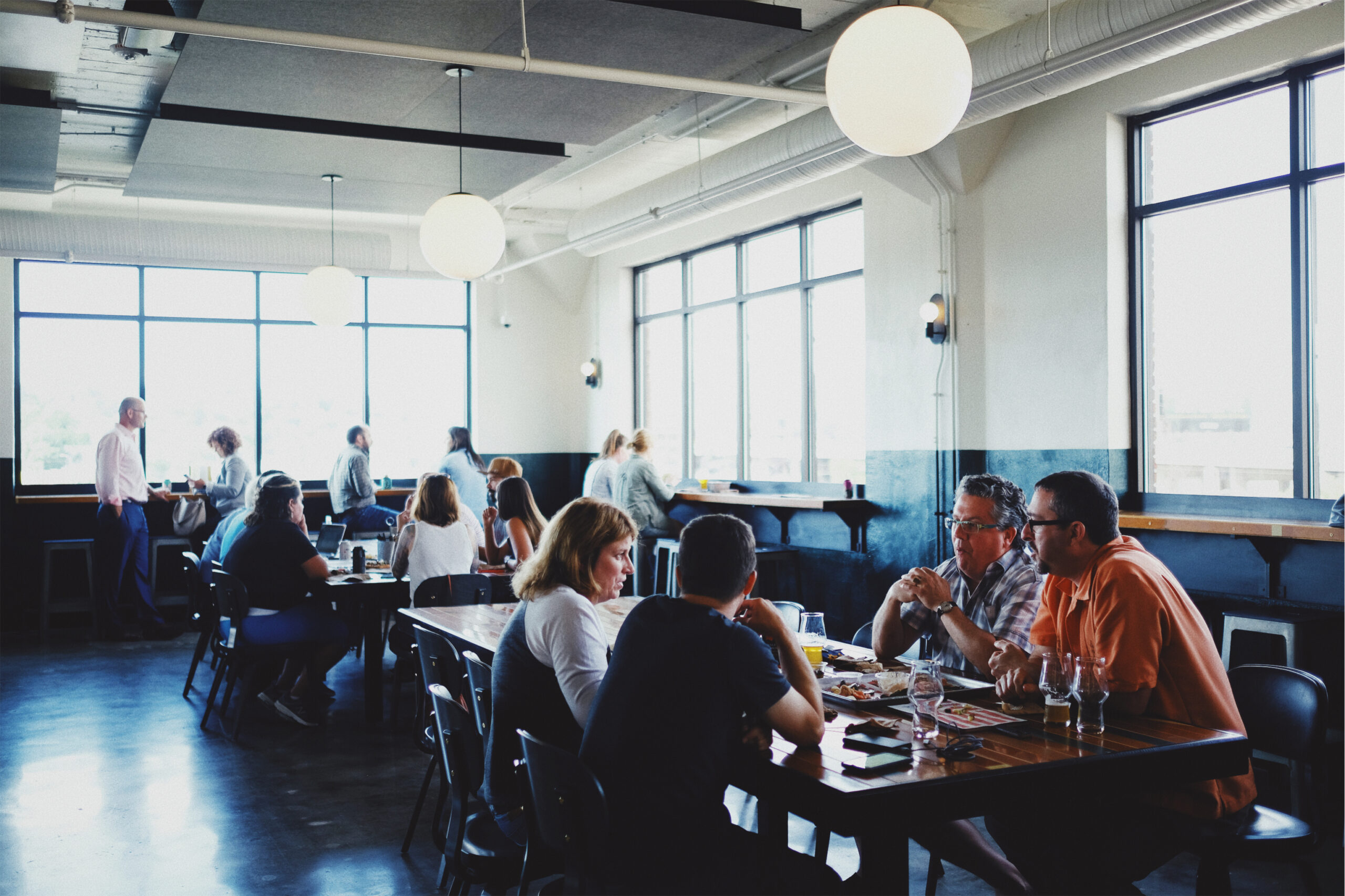 A group of people is sitting at a wooden table, engaging in conversation inside a well-lit, modern cafe with large windows. In the background, other patrons are seated and standing near the windows. The room features high ceilings and hanging globe lights.